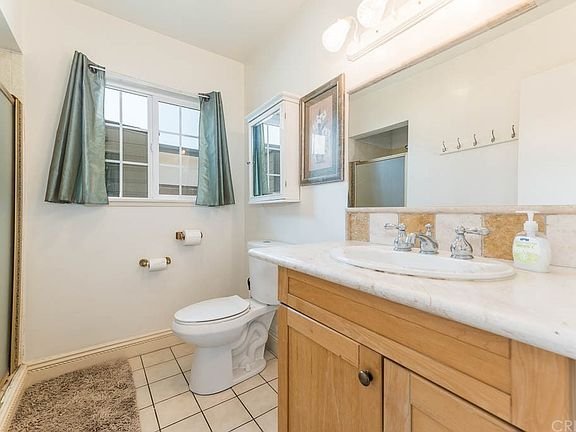 California bathroom with wooden vanity, marble countertop, mirror, toilet, and window with green curtains
